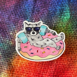 May include: A raccoon wearing sunglasses and a life preserver floats in a pink donut with sprinkles. The donut is on a tie-dye background.