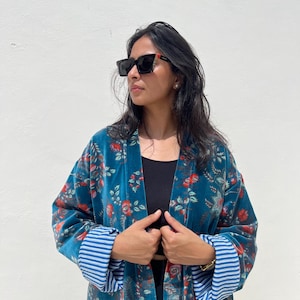 May include: A teal velvet kimono with a floral pattern in red and white. The kimono has blue and white striped cuffs. The person in the image is wearing black sunglasses and a black top.