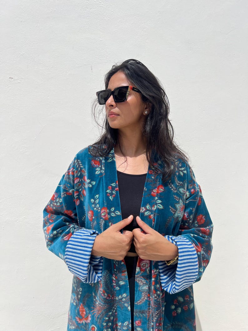 May include: A teal velvet kimono with a floral pattern in red and white. The kimono has blue and white striped cuffs. The person in the image is wearing black sunglasses and a black top.