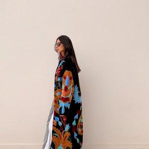 May include: A long black coat with colourful floral embroidery in shades of orange, blue, and red. The coat is worn over striped trousers and a white top. The person is wearing sunglasses and brown boots.