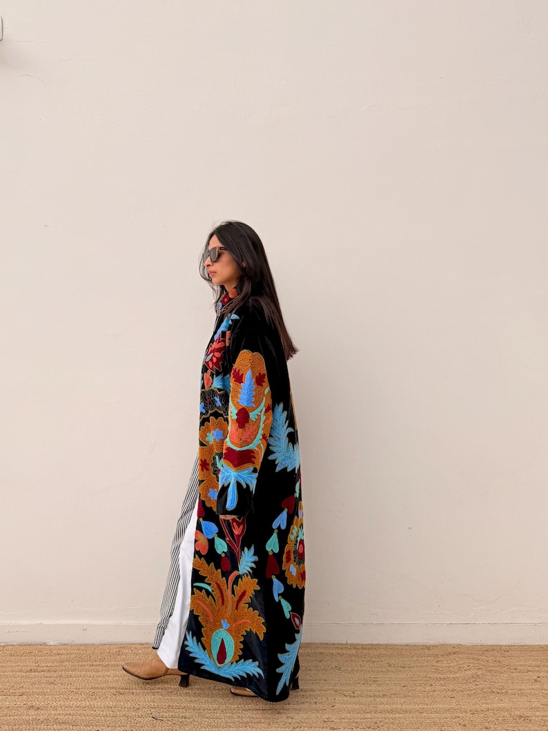 May include: A long black coat with colourful floral embroidery in shades of orange, blue, and red. The coat is worn over striped trousers and a white top. The person is wearing sunglasses and brown boots.
