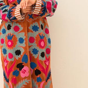 Puede incluir: Abrigo largo de color &oacute;xido con intrincados bordados florales en tonos rosa, azul y verde. El abrigo presenta una textura aterciopelada y un patr&oacute;n detallado. Las mangas tienen pu&ntilde;os a rayas.