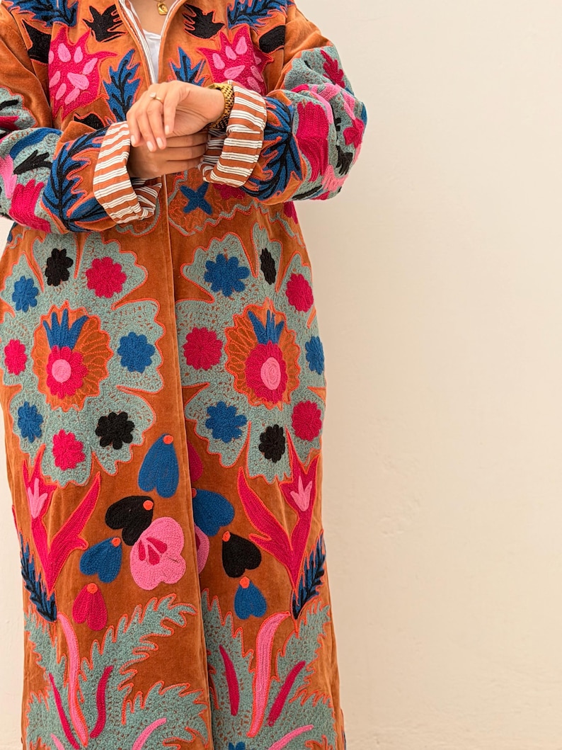 Puede incluir: Abrigo largo de color &oacute;xido con intrincados bordados florales en tonos rosa, azul y verde. El abrigo presenta una textura aterciopelada y un patr&oacute;n detallado. Las mangas tienen pu&ntilde;os a rayas.