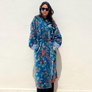 May include: A long, teal velvet robe with a floral pattern in red and orange. The robe has a matching belt and striped cuffs. The person is wearing black sunglasses, black pants, and tan boots.