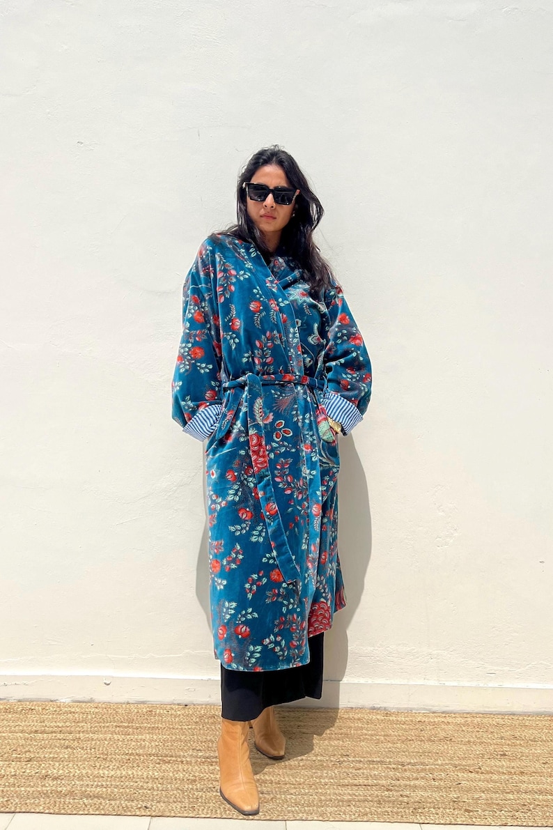 May include: A long, teal velvet robe with a floral pattern in red and orange. The robe has a matching belt and striped cuffs. The person is wearing black sunglasses, black pants, and tan boots.