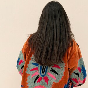 Puede incluir: Chaqueta de terciopelo naranja con intrincados bordados florales en tonos azules, rosas y verdes. La chaqueta presenta un gran dise&ntilde;o bordado en la espalda y mangas, de estilo bohemio.