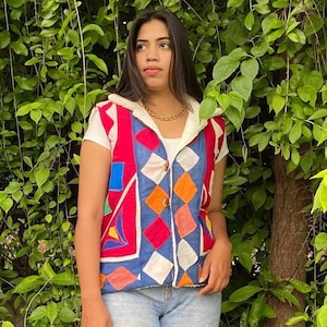 May include: A colorful, quilted vest with a geometric pattern. The vest features red, blue, orange, and white shapes, with a cream-colored, hooded lining. It has a button closure and is worn over a white t-shirt and blue jeans.