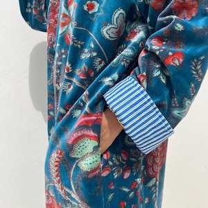 May include: A teal velvet robe with a floral pattern in red, pink, and green. The robe features a pocket and blue and white striped cuffs. The fabric has a soft, luxurious appearance.
