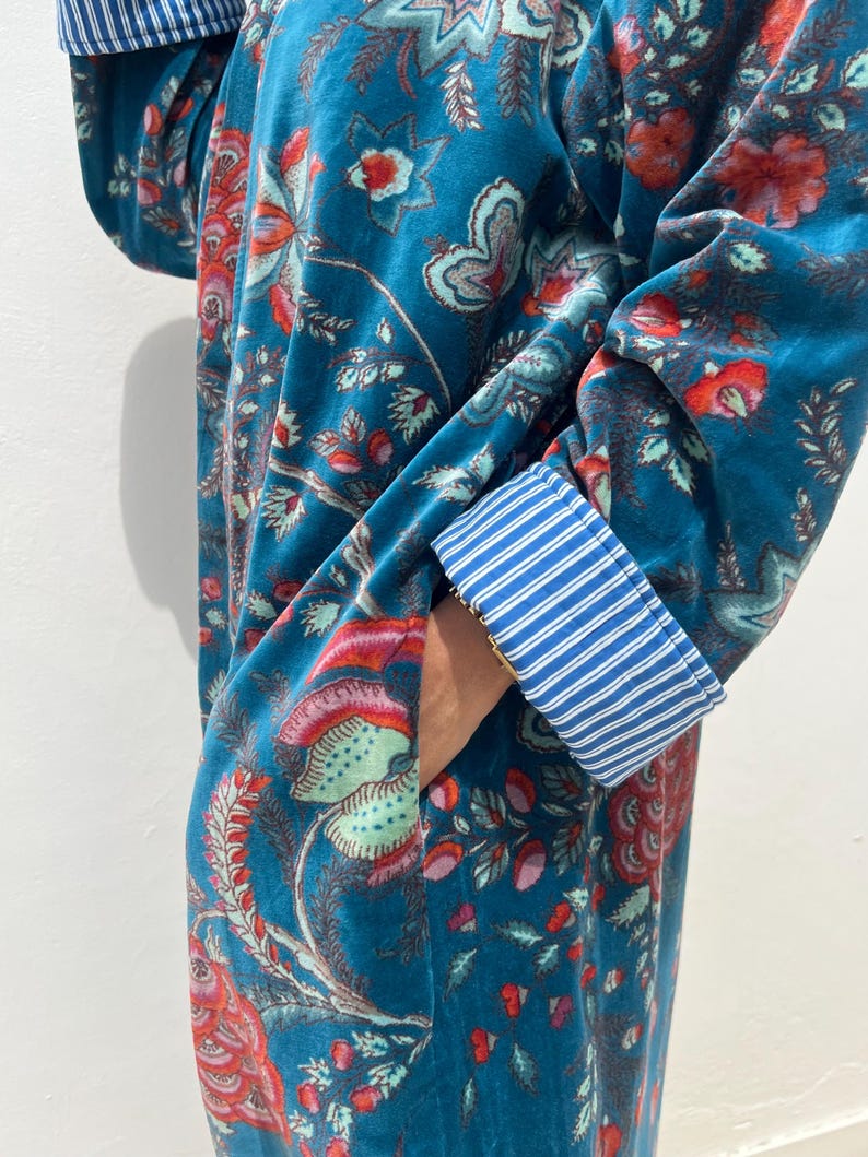 May include: A teal velvet robe with a floral pattern in red, pink, and green. The robe features a pocket and blue and white striped cuffs. The fabric has a soft, luxurious appearance.