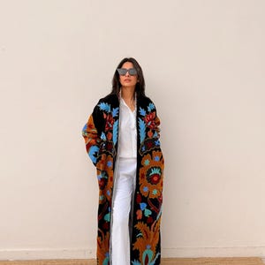 May include: A long, black embroidered coat with colourful floral patterns in shades of orange, blue, and green. The coat is worn over a white shirt and trousers. The coat's design is a striking visual element.