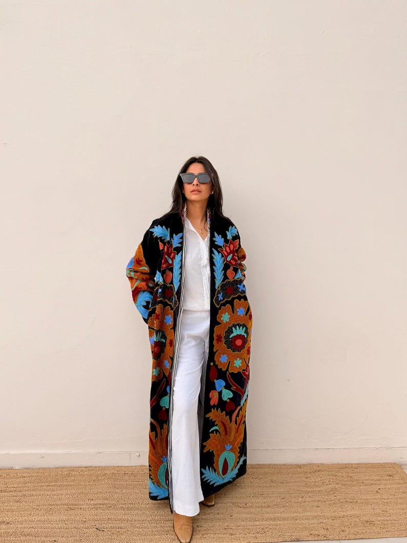 May include: A long, black embroidered coat with colourful floral patterns in shades of orange, blue, and green. The coat is worn over a white shirt and trousers. The coat's design is a striking visual element.