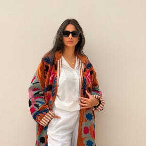 Puede incluir: Un abrigo largo de color &oacute;xido con bordados florales coloridos. El abrigo presenta un forro blanco y pu&ntilde;os a rayas. La persona de la imagen lleva gafas de sol negras, una camisa blanca y pantalones blancos. El abrigo es una pieza llamativa.