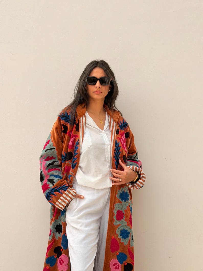 Puede incluir: Un abrigo largo de color &oacute;xido con bordados florales coloridos. El abrigo presenta un forro blanco y pu&ntilde;os a rayas. La persona de la imagen lleva gafas de sol negras, una camisa blanca y pantalones blancos. El abrigo es una pieza llamativa.