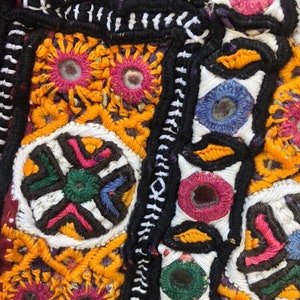 Puede incluir: Primer plano de un textil colorido con intrincados bordados. El dise&ntilde;o presenta formas geom&eacute;tricas, motivos florales y peque&ntilde;os espejos. Los colores predominantes incluyen amarillo, naranja, rosa, verde, azul y negro, creando una superficie vibrante y detallada.