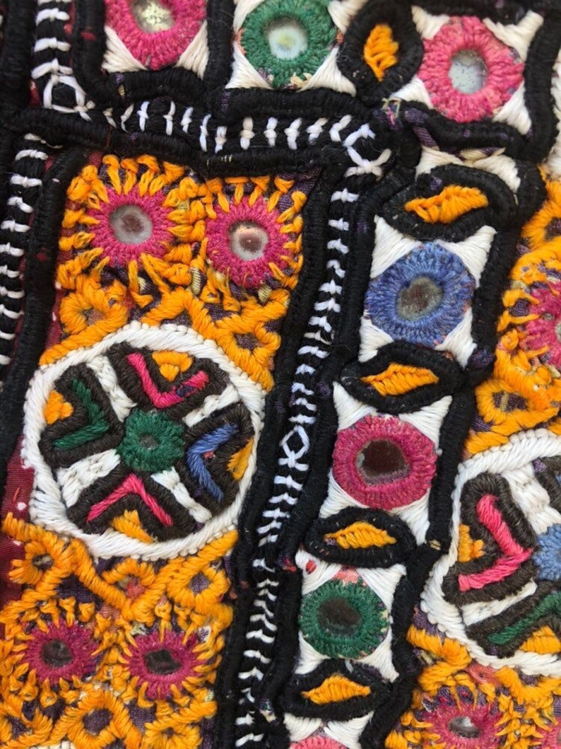 Puede incluir: Primer plano de un textil colorido con intrincados bordados. El dise&ntilde;o presenta formas geom&eacute;tricas, motivos florales y peque&ntilde;os espejos. Los colores predominantes incluyen amarillo, naranja, rosa, verde, azul y negro, creando una superficie vibrante y detallada.