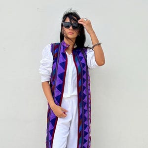 May include: A woman wearing a white outfit with a long, sleeveless vest. The vest is a vibrant purple with geometric patterns in shades of blue, red, and teal. She is also wearing black sunglasses.