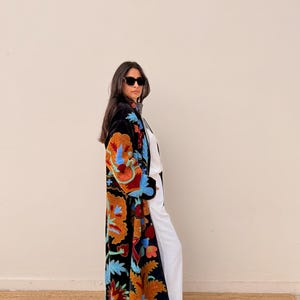 May include: A long, black embroidered coat with vibrant floral patterns in orange, blue, and red. The coat is worn over white trousers and a white top. The coat's design features intricate details and a flowing silhouette, suitable for a variety of occasions.
