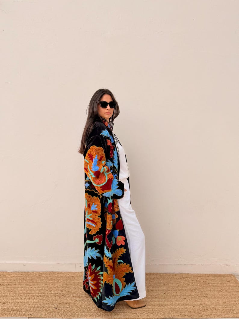 May include: A long, black embroidered coat with vibrant floral patterns in orange, blue, and red. The coat is worn over white trousers and a white top. The coat's design features intricate details and a flowing silhouette, suitable for a variety of occasions.
