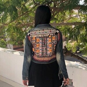 Puede incluir: Chaqueta vaquera con intrincados bordados y adornos en la espalda. La chaqueta presenta un denim de lavado oscuro con patrones coloridos y detalles met&aacute;licos. La persona lleva una falda negra.