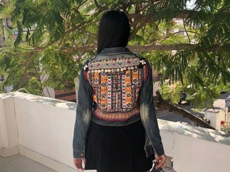 Puede incluir: Chaqueta vaquera con intrincados bordados y adornos en la espalda. La chaqueta presenta un denim de lavado oscuro con patrones coloridos y detalles met&aacute;licos. La persona lleva una falda negra.