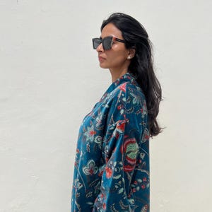 May include: A teal floral patterned robe with a blue and white striped cuff. The robe is made of a soft, velvety material. The person is wearing black sunglasses.