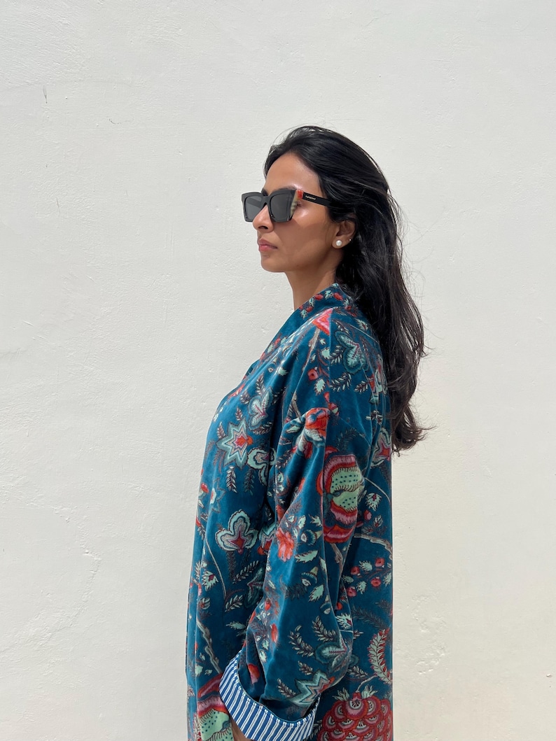 May include: A teal floral patterned robe with a blue and white striped cuff. The robe is made of a soft, velvety material. The person is wearing black sunglasses.