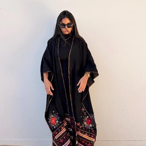 May include: A black embroidered kimono-style garment with gold trim and colorful floral and geometric patterns at the hem. The garment is worn over a black outfit. The image is taken in front of a white wall.