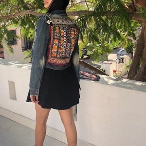 Puede incluir: Chaqueta vaquera con un panel trasero bordado con un dise&ntilde;o geom&eacute;trico colorido. La chaqueta se combina con una falda negra y botines negros. La imagen est&aacute; tomada al aire libre con un &aacute;rbol al fondo.