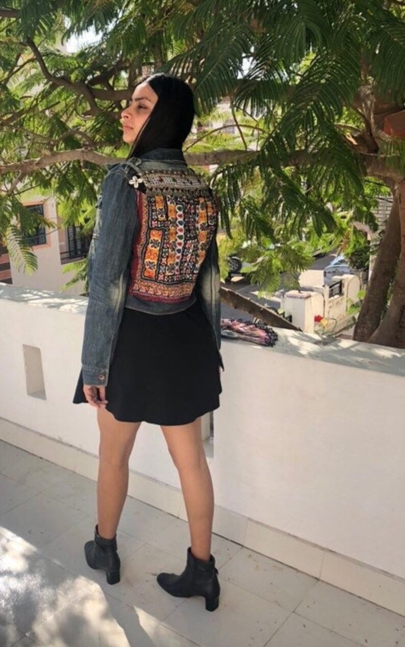 Puede incluir: Chaqueta vaquera con un panel trasero bordado con un dise&ntilde;o geom&eacute;trico colorido. La chaqueta se combina con una falda negra y botines negros. La imagen est&aacute; tomada al aire libre con un &aacute;rbol al fondo.