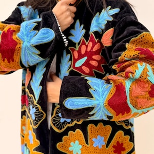 May include: Black velvet coat with colourful floral embroidery in blue, red, orange, and yellow. The coat has long sleeves and a mandarin collar. The embroidery is detailed and covers the sleeves and front panels.