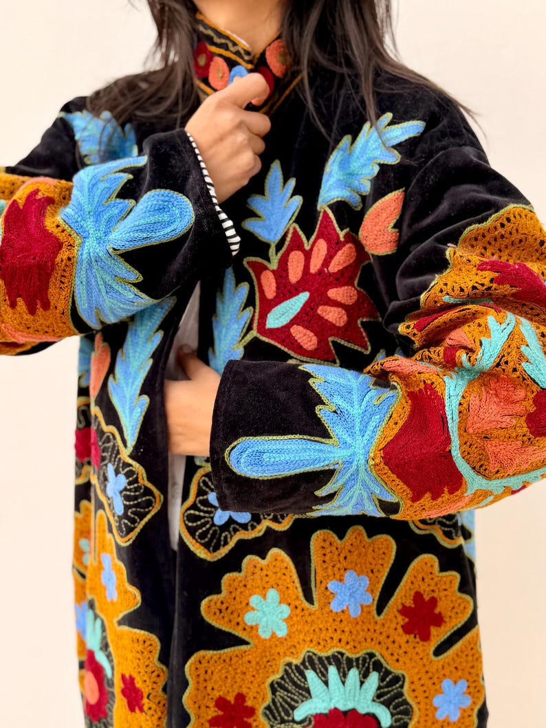 May include: Black velvet coat with colourful floral embroidery in blue, red, orange, and yellow. The coat has long sleeves and a mandarin collar. The embroidery is detailed and covers the sleeves and front panels.