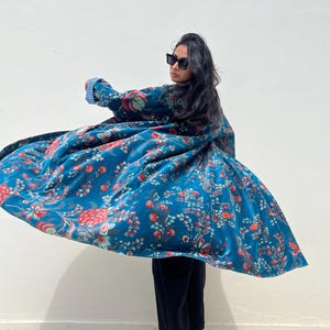 May include: A flowing, teal-colored coat with a floral pattern in red, pink, and green. The coat is made of a soft, velvety material. The sleeves have a blue and white striped lining. The person is wearing black pants and tan boots.