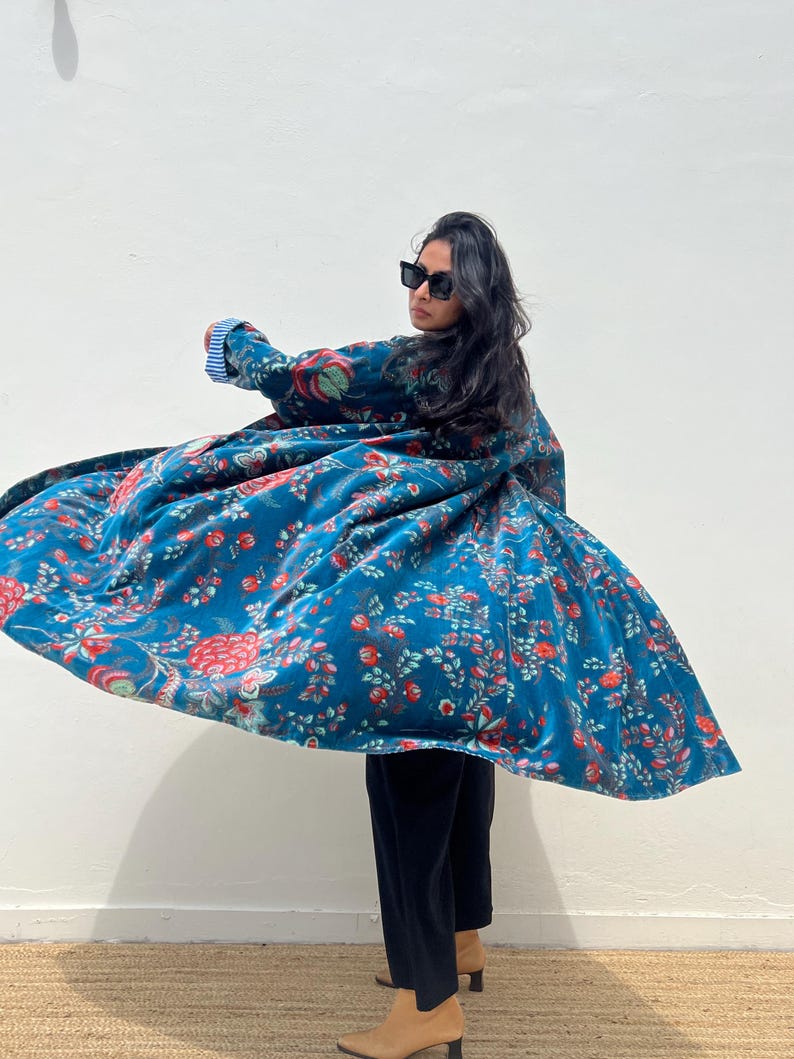 May include: A flowing, teal-colored coat with a floral pattern in red, pink, and green. The coat is made of a soft, velvety material. The sleeves have a blue and white striped lining. The person is wearing black pants and tan boots.