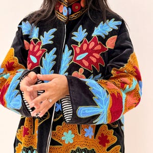 May include: Black velvet coat with colourful floral embroidery in red, blue, and yellow. The coat has a high collar and a full-length zip. The sleeves have a striped lining. A stylish outerwear piece.