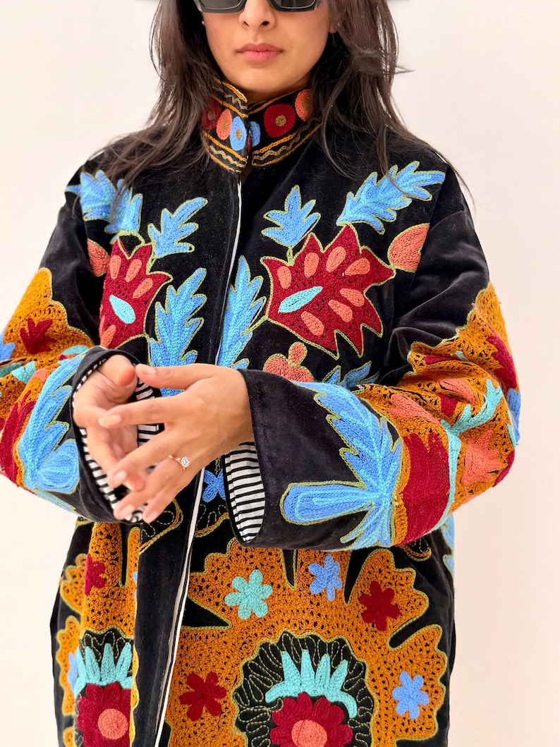May include: Black velvet coat with colourful floral embroidery in red, blue, and yellow. The coat has a high collar and a full-length zip. The sleeves have a striped lining. A stylish outerwear piece.
