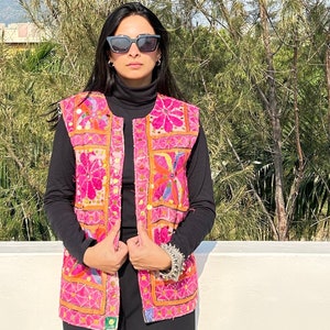 Peut inclure: Un gilet brodé rose avec des motifs floraux colorés et des embellissements en miroir. Le gilet a une fermeture à boutons et un fond noir.