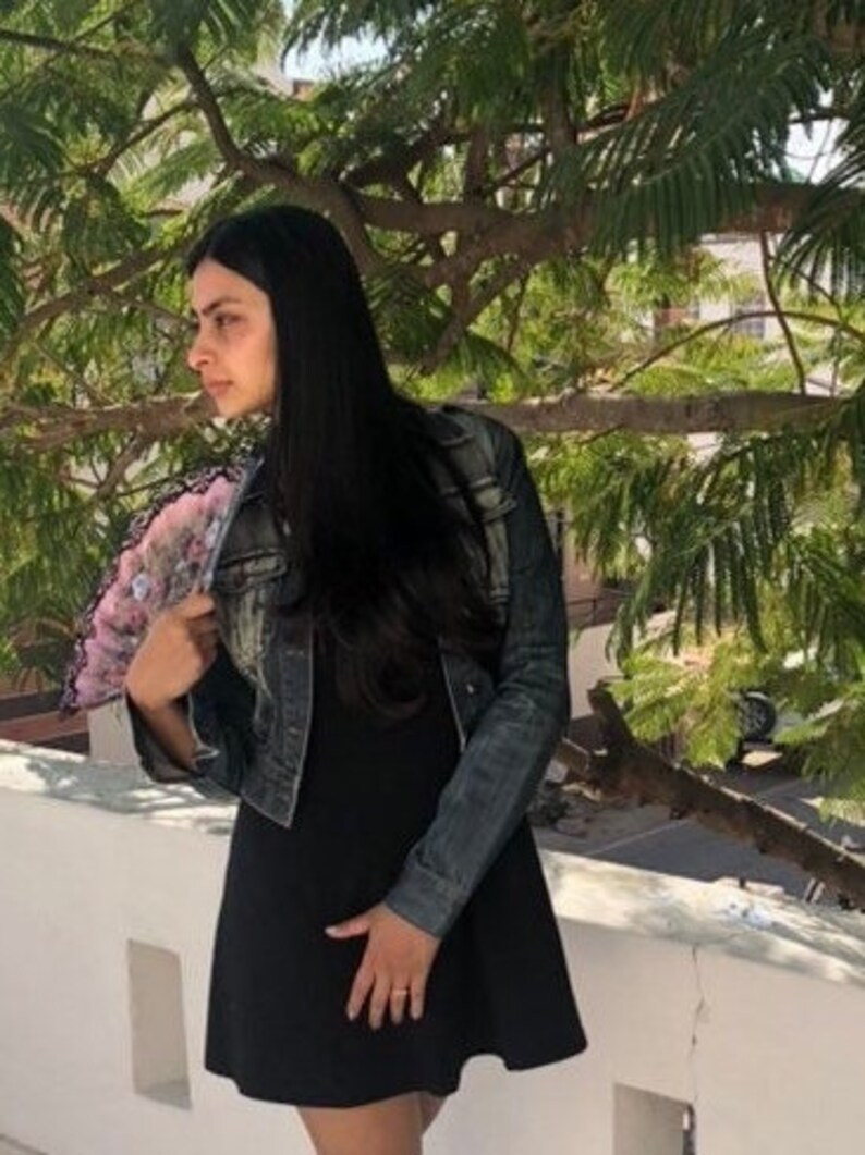 Puede incluir: Un vestido negro y una chaqueta vaquera. El vestido es de dise&ntilde;o sencillo y sin mangas. La chaqueta es de estilo vaquero cl&aacute;sico, con un lavado descolorido. La persona sostiene un abanico decorativo con motivos florales rosas y blancos.