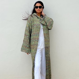 May include: A long, hooded coat in a speckled green, brown, and red knit fabric. The coat is worn over a white top and pants. The coat has a button closure and wide sleeves. The person is wearing black sunglasses.