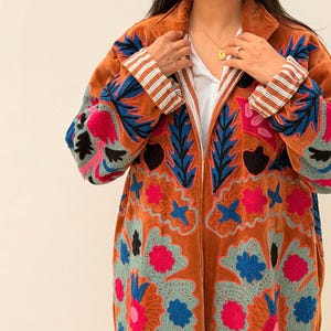 Puede incluir: Un abrigo largo de color &oacute;xido con intrincados bordados florales en tonos azules, rosas y turquesas. El abrigo presenta un cuello con solapas y pu&ntilde;os a rayas. El bordado crea un contraste llamativo sobre el fondo c&aacute;lido.