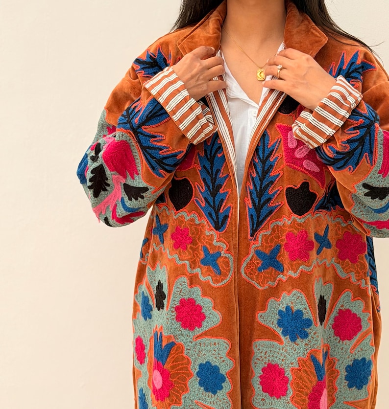 Puede incluir: Un abrigo largo de color &oacute;xido con intrincados bordados florales en tonos azules, rosas y turquesas. El abrigo presenta un cuello con solapas y pu&ntilde;os a rayas. El bordado crea un contraste llamativo sobre el fondo c&aacute;lido.