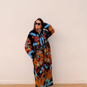 May include: A long, black embroidered coat with a vibrant floral design in shades of orange, red, blue, and teal. The coat features a full-length zip and is worn by a person wearing sunglasses.