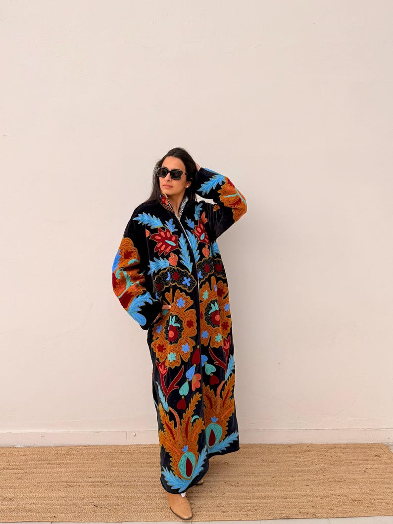 May include: A long, black embroidered coat with a vibrant floral design in shades of orange, red, blue, and teal. The coat features a full-length zip and is worn by a person wearing sunglasses.