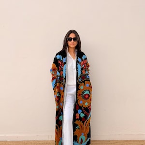 May include: A long, black coat featuring an elaborate floral embroidery in blue, orange, and brown. The coat is worn over a white shirt and trousers. The detailed pattern of the coat provides a bohemian aesthetic to the ensemble.