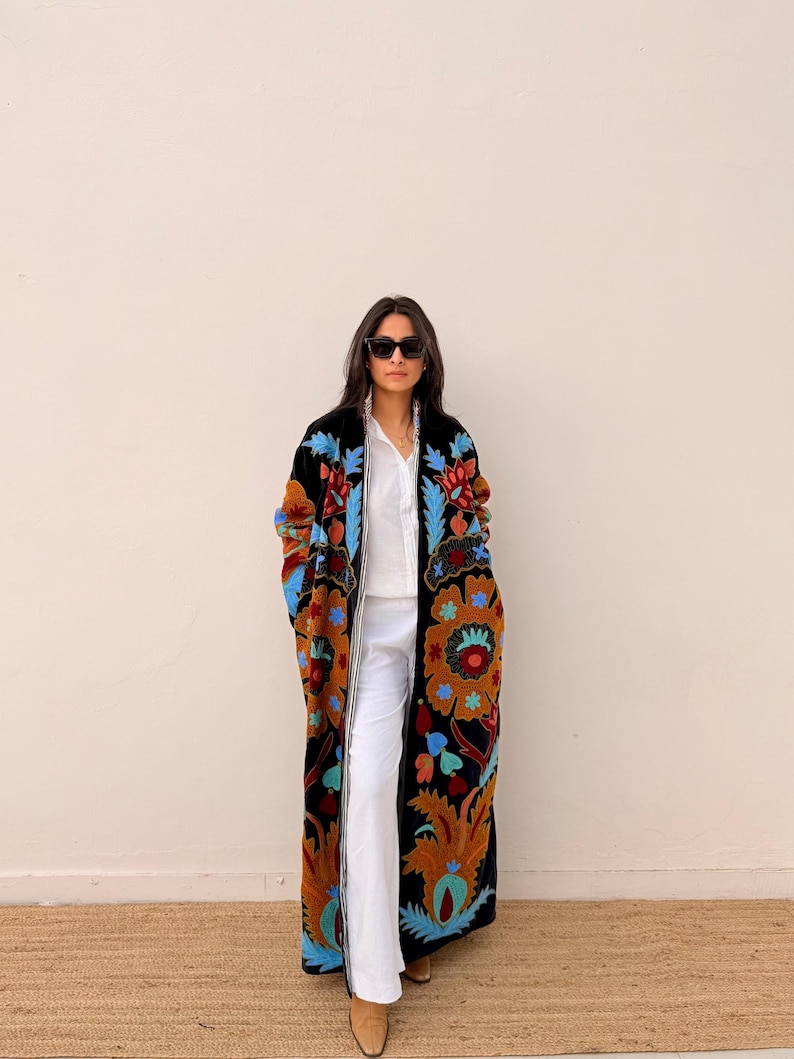May include: A long, black coat featuring an elaborate floral embroidery in blue, orange, and brown. The coat is worn over a white shirt and trousers. The detailed pattern of the coat provides a bohemian aesthetic to the ensemble.