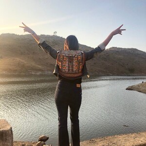 Puede incluir: Una persona con una chaqueta vaquera con un panel trasero bordado, pantalones negros y botas, de pie con los brazos extendidos cerca de un lago. La chaqueta presenta un patr&oacute;n colorido e intrincado. La escena se desarrolla contra un tel&oacute;n de fondo de un lago y colinas bajo un cielo despejado.