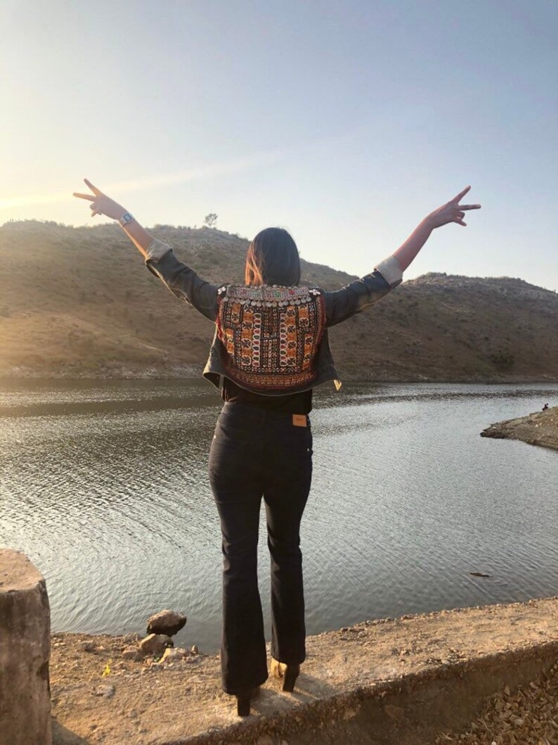 Puede incluir: Una persona con una chaqueta vaquera con un panel trasero bordado, pantalones negros y botas, de pie con los brazos extendidos cerca de un lago. La chaqueta presenta un patr&oacute;n colorido e intrincado. La escena se desarrolla contra un tel&oacute;n de fondo de un lago y colinas bajo un cielo despejado.