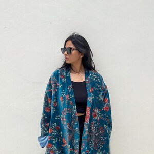 May include: A teal velvet kimono with a floral pattern in red and green. The kimono has a relaxed fit and long sleeves. The cuffs are lined with a blue and white striped fabric. The person is wearing black sunglasses.