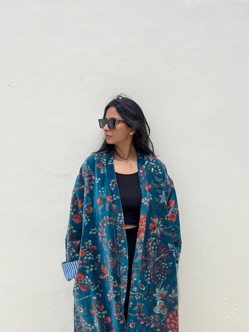May include: A teal velvet kimono with a floral pattern in red and green. The kimono has a relaxed fit and long sleeves. The cuffs are lined with a blue and white striped fabric. The person is wearing black sunglasses.