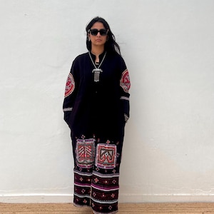 May include: A black long dress with colorful embroidered details on the sleeves and lower portion. The dress features red, pink, and white geometric patterns. The person is wearing sunglasses and a silver necklace. The dress is worn with tan boots.