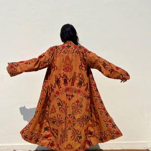 May include: A long, rust-colored robe with a floral and paisley pattern. The robe has a mandarin collar and long sleeves. The robe is embroidered with figures and text in black, red, and white.